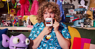 Business owner Rachel is sitting on a couch with her purple puppet next to her, holding a branded 'Rachel Made Studio' mug up to her mouth